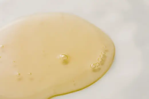 Ein Tropfen klares Pflegeöl auf heller Fläche, im Gegenlicht fotografiert.
